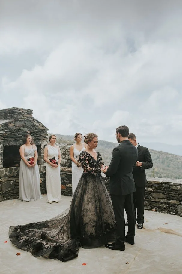 Outdoor mountain ceremony with the bride in a black wedding dress and the wedding party standing near a stone wall.