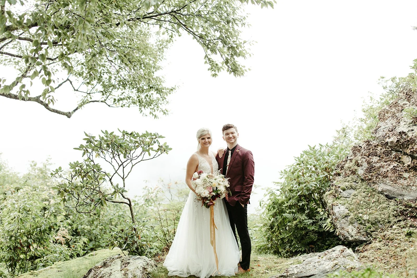 Bride and groom smiling in a lush mountain forest setting, bride holding a bouquet with long ribbon