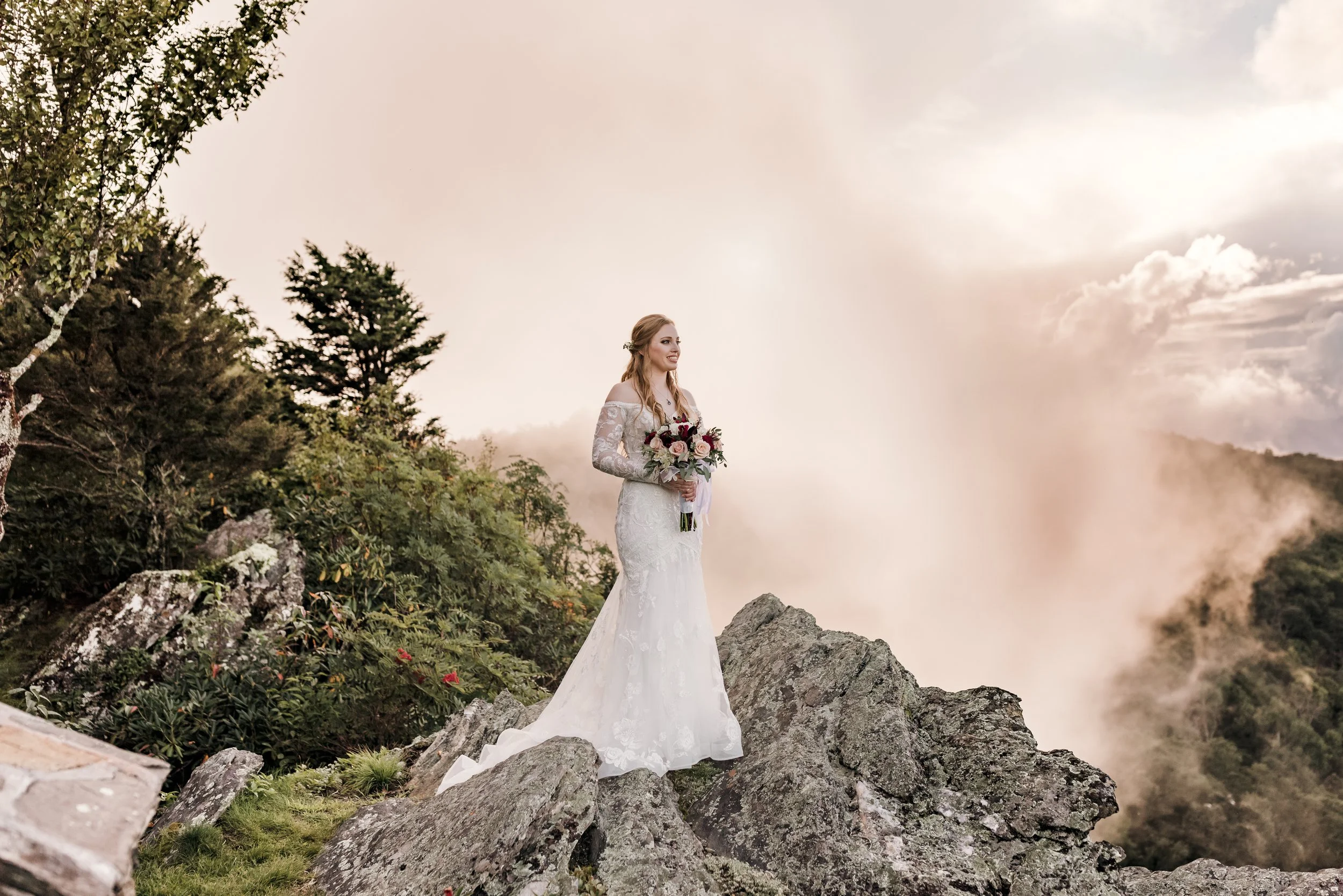 Bride in an off-the-shoulder lace wedding dress standing on rocky mountaintop with misty mountains at sunset, holding a bouquet.