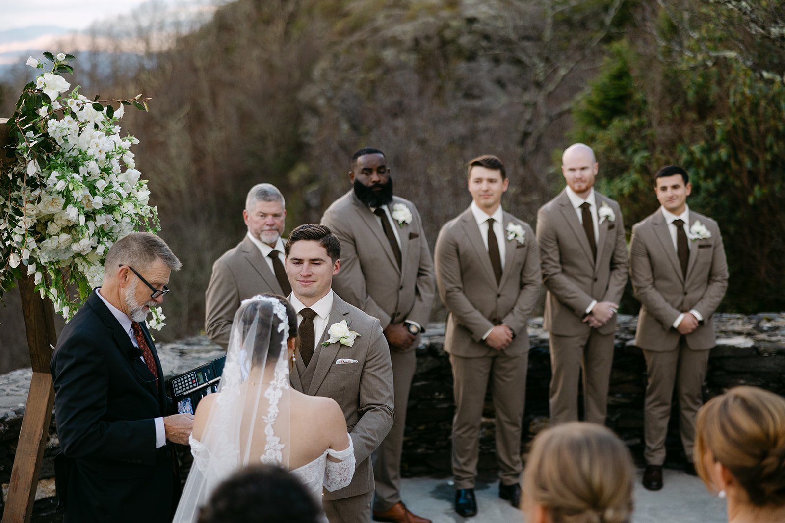 Groom and groomsmen in taupe suits standing during an outdoor mountain wedding ceremony with a white floral arch.