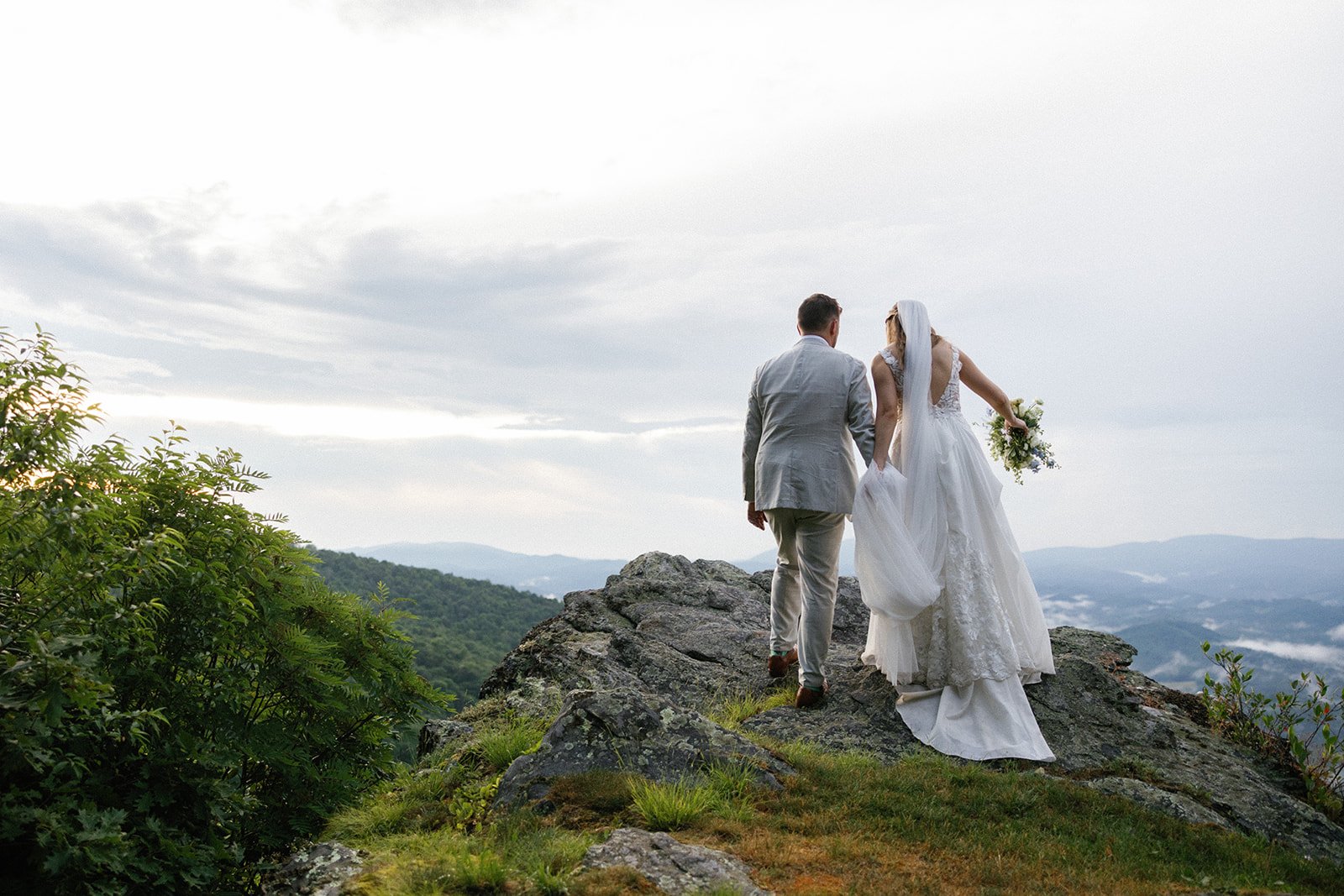 Bride and groom standing together on a rocky mountain overlook with misty blue ridge views.