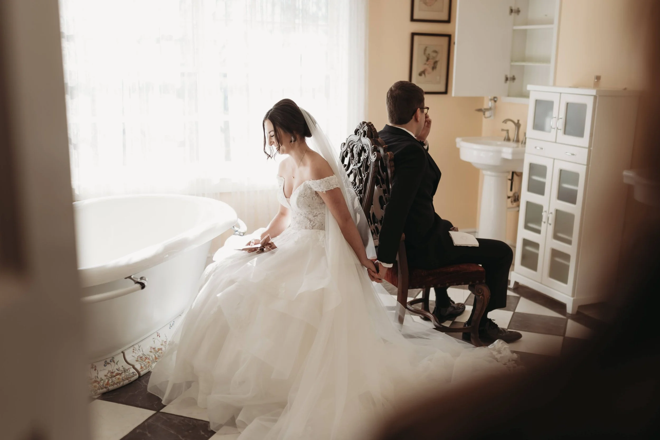 Bride and groom sitting back to back holding hands during a first look in a bright bathroom.