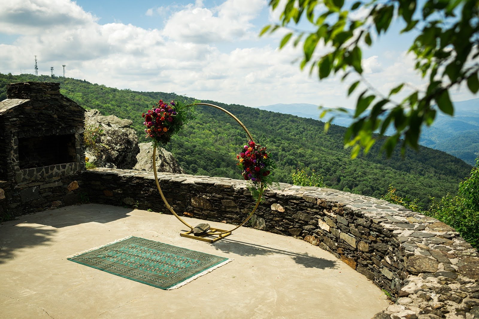Outdoor ceremony space with a gold circular arch decorated with flowers, set on a stone terrace overlooking green mountains.