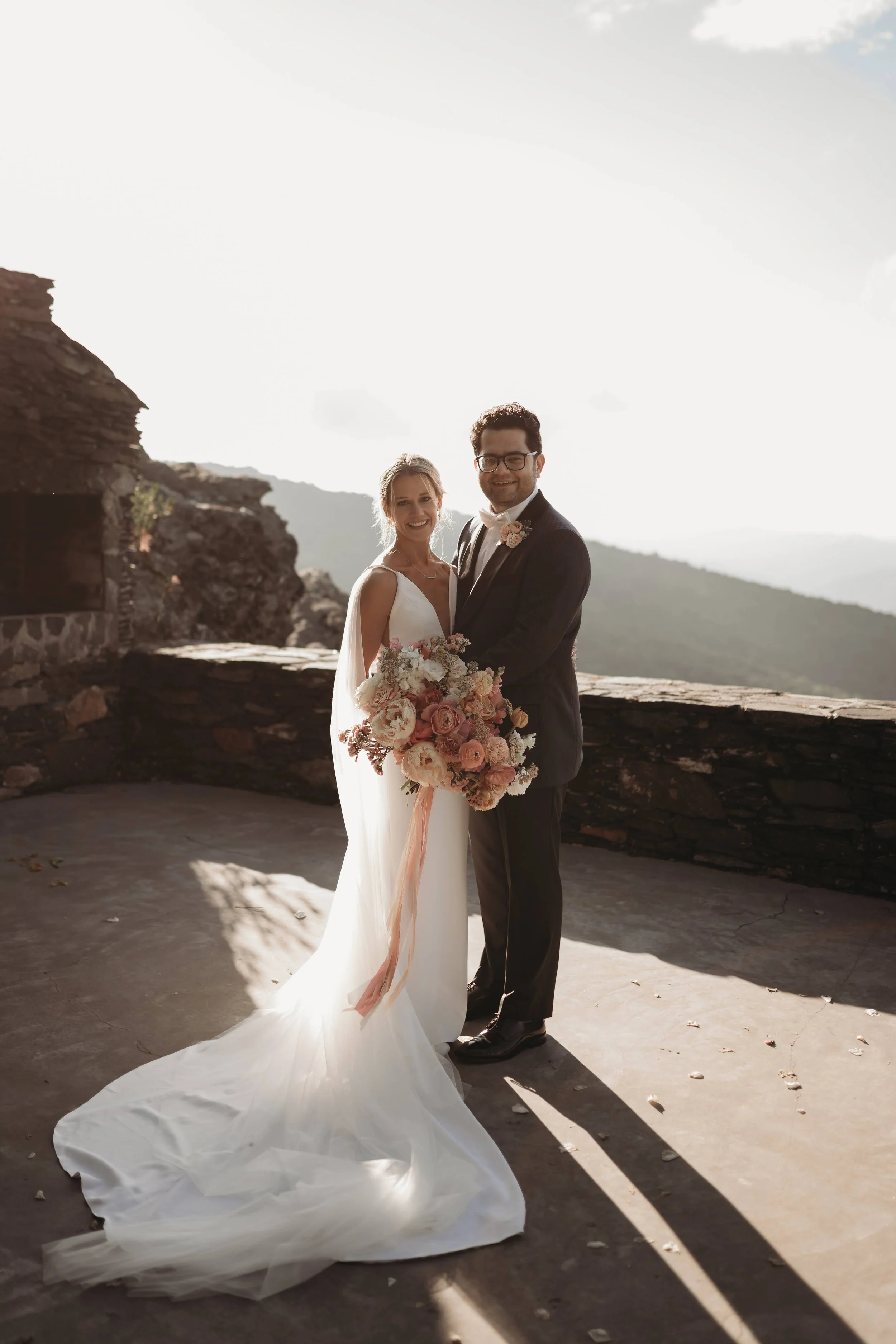 Bride and groom standing together at a sunny mountain overlook with a blush bouquet and stone wall behind them.