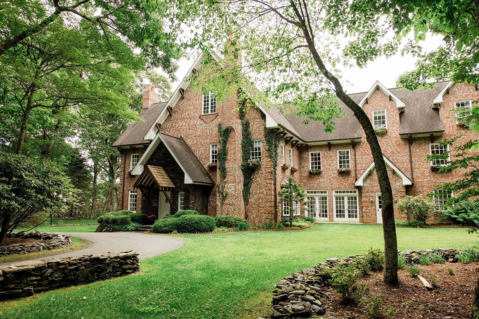 Historic brick estate wedding venue exterior surrounded by trees, stone walls, manicured lawn, and lush greenery.
