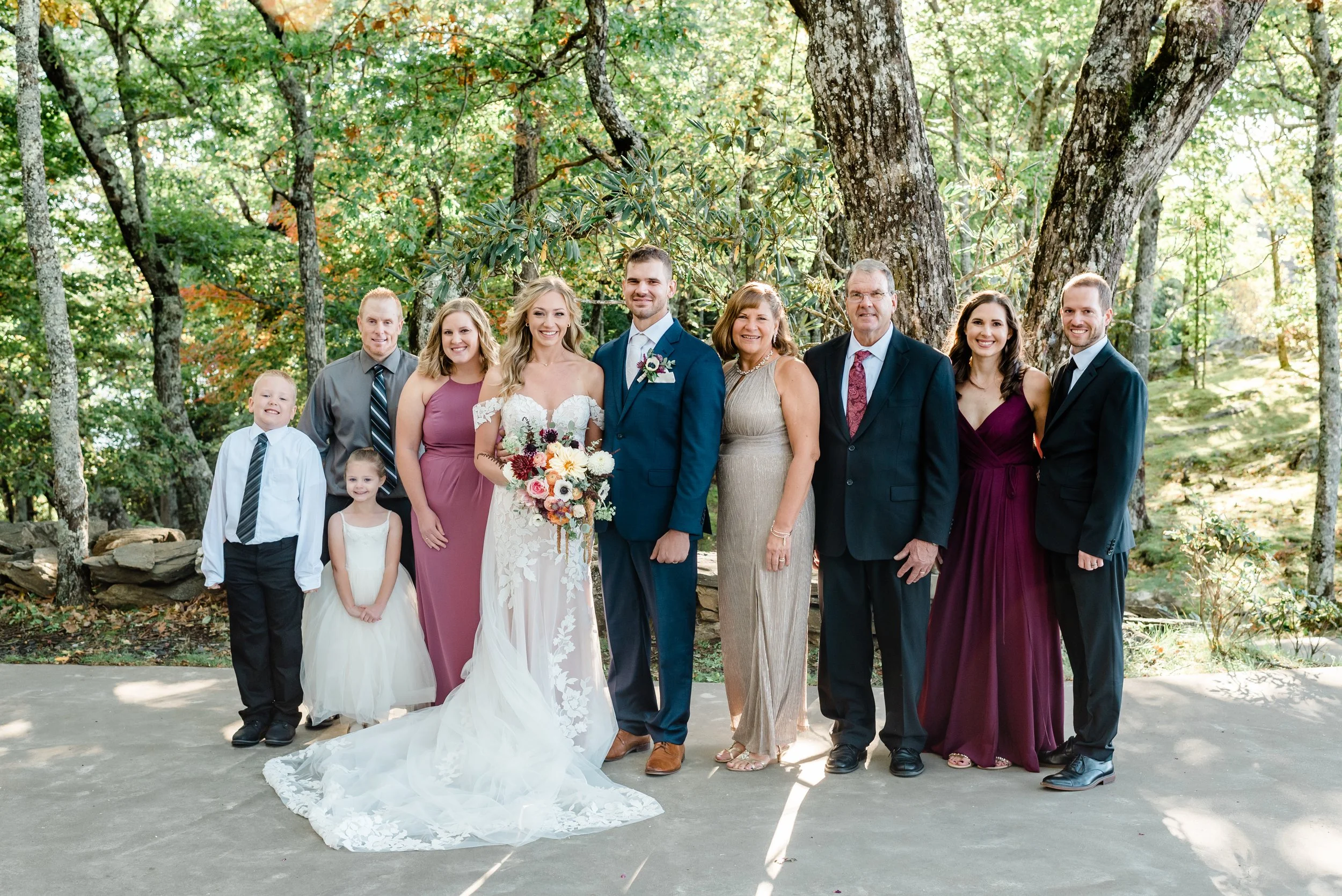 Bride and groom posing with family members in a wooded outdoor setting with autumn florals and formal wedding attire.