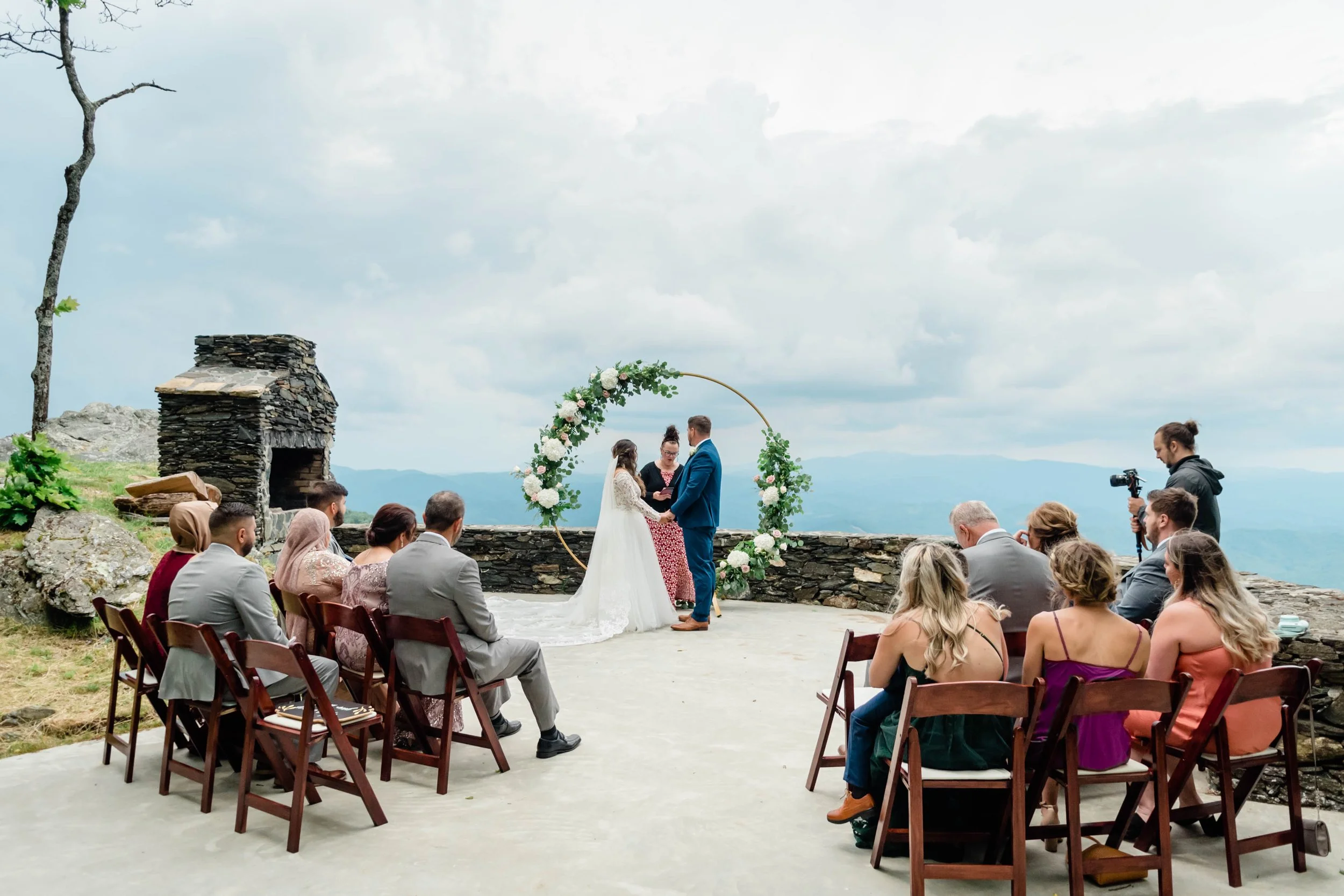 Wide view of a mountaintop wedding ceremony with guests seated on wooden chairs and a floral arch at the front