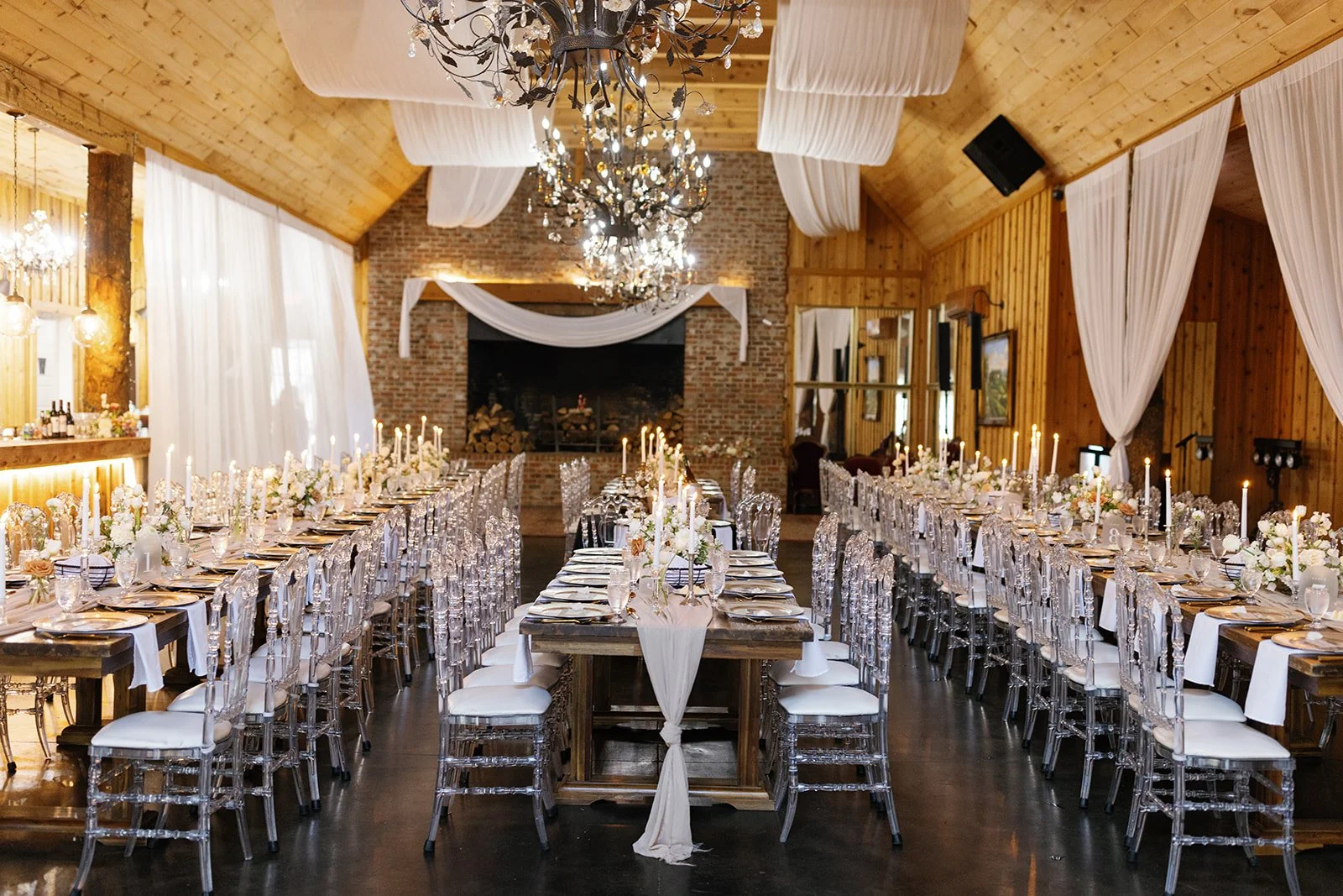Long head table with clear chairs and candlelight set in a wooden reception hall with chandeliers and draped fabric