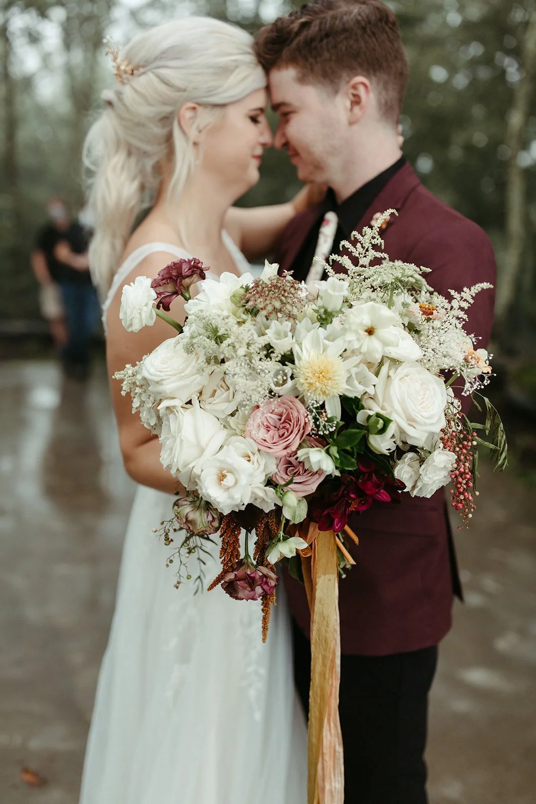Bride and groom embracing closely with a lush white, blush, and burgundy floral bouquet in the foreground.
