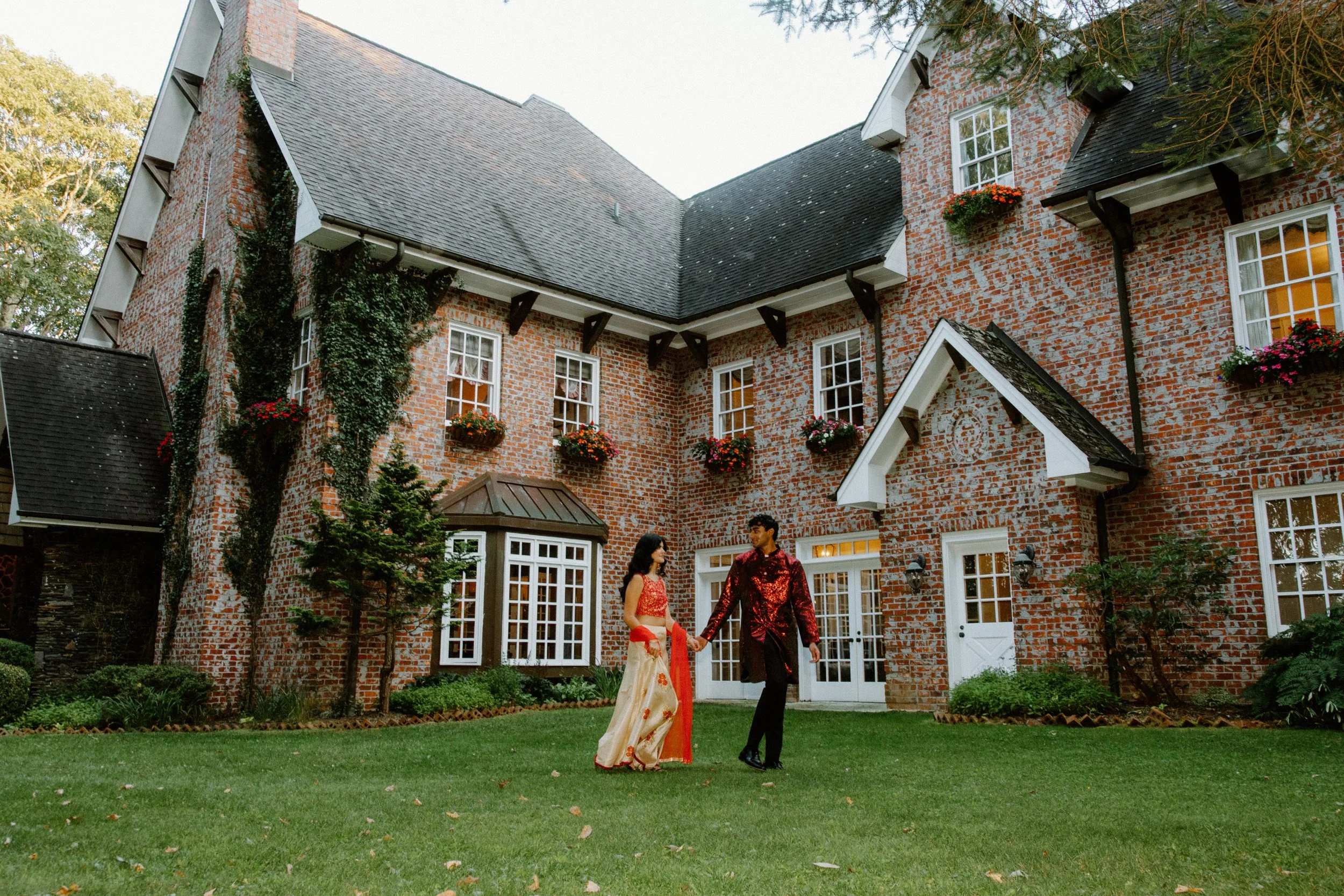 Couple walks hand-in-hand on a lawn in front of a large ivy-covered brick manor with flower boxes and white doors.