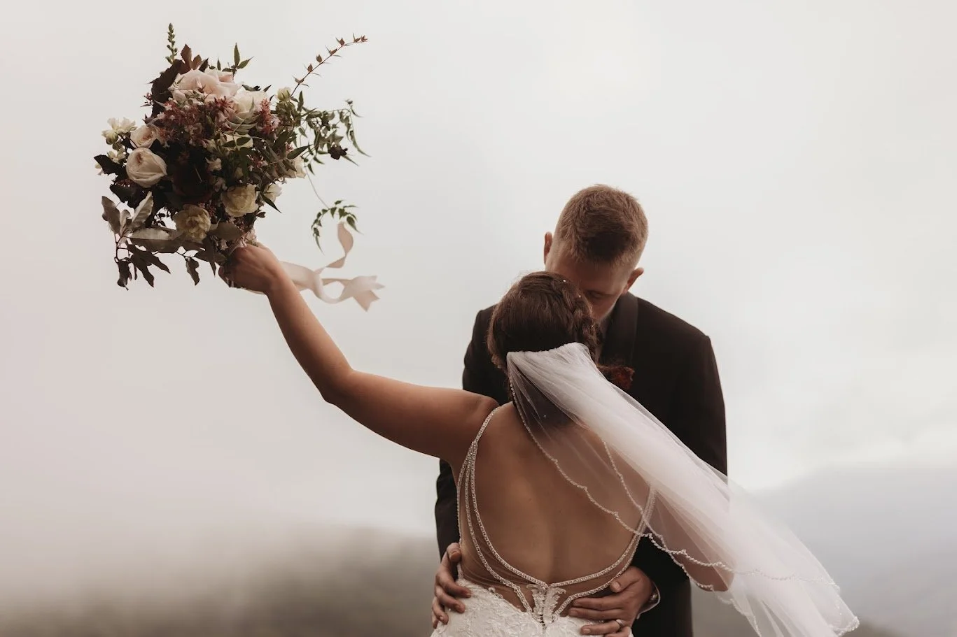 Bride and groom kissing as the bride lifts her bouquet, with foggy mountain views behind them