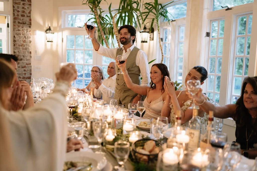 Groom raising a wine glass during a candlelit estate wedding reception while guests applaud around the table.
