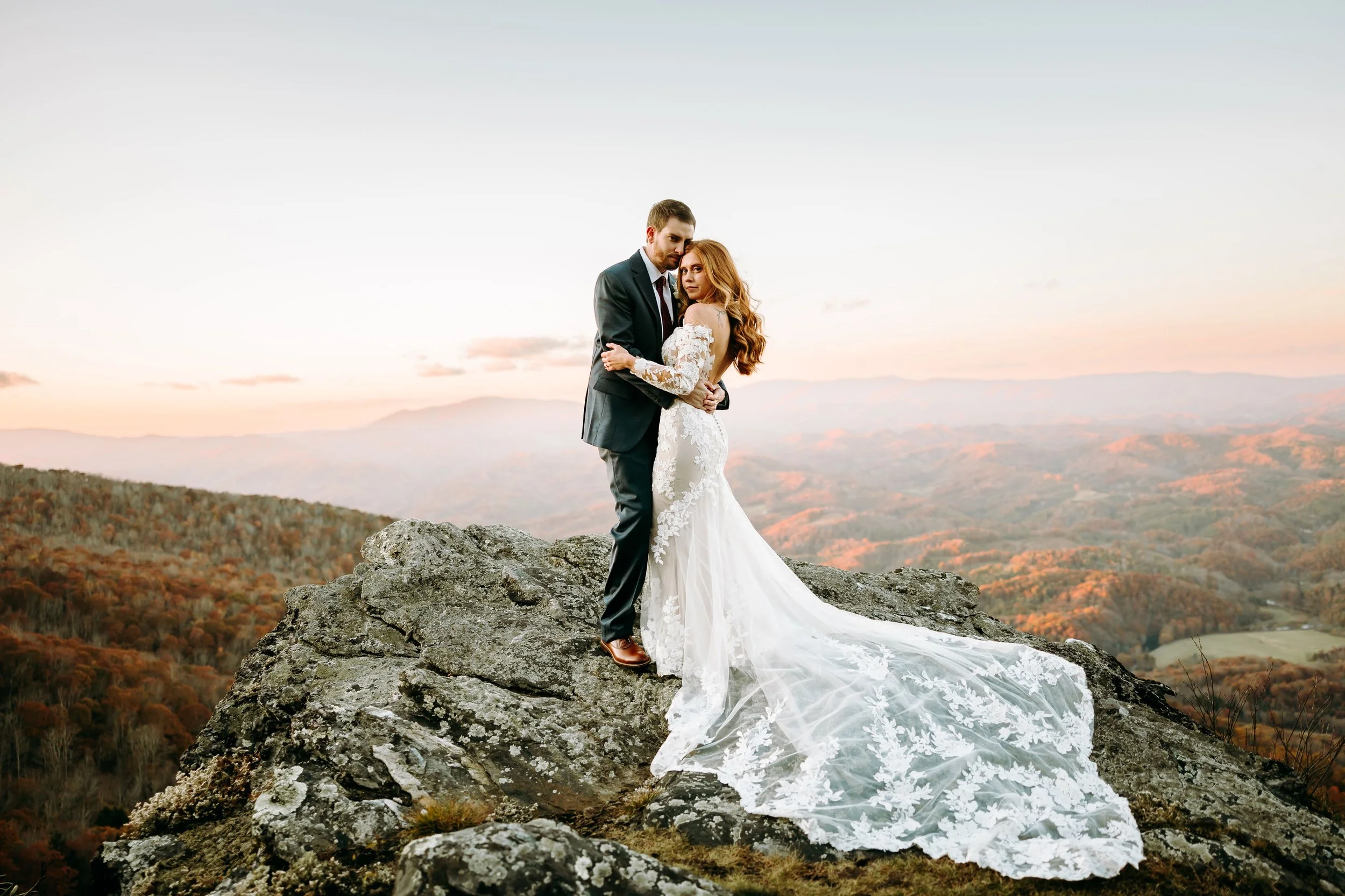 Bride and groom posing on a rocky cliff at sunset with a sweeping mountain landscape