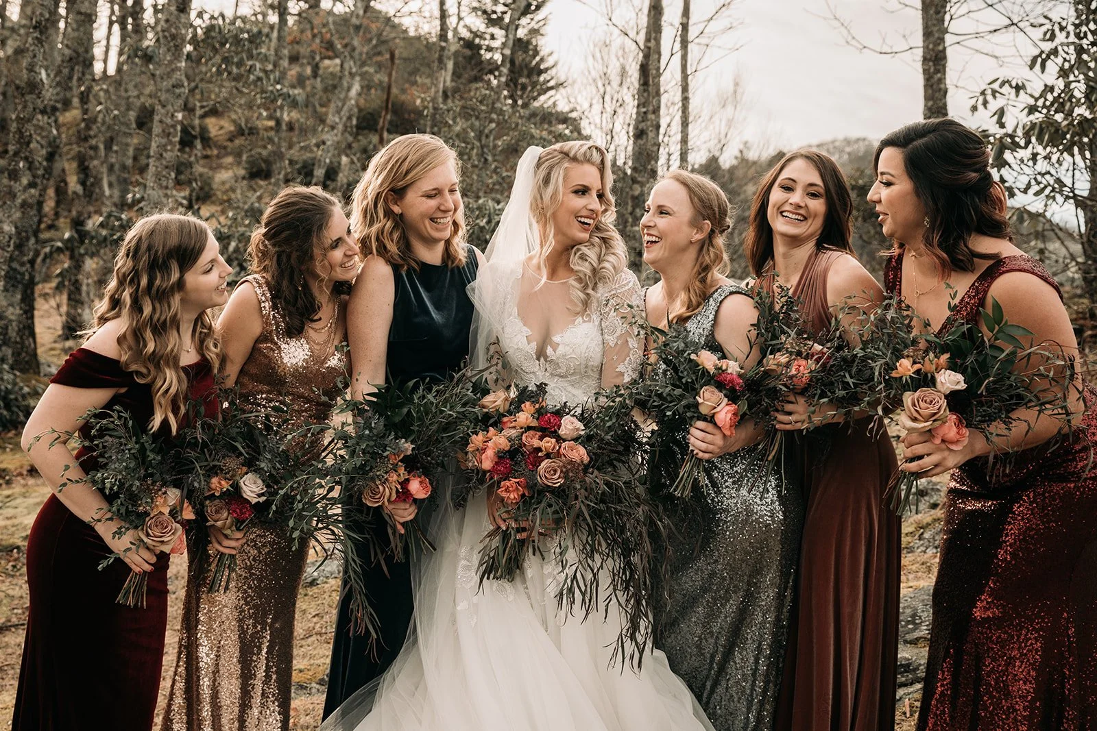 Bride laughing with bridesmaids holding bouquets, wearing mixed deep-toned dresses outdoors.