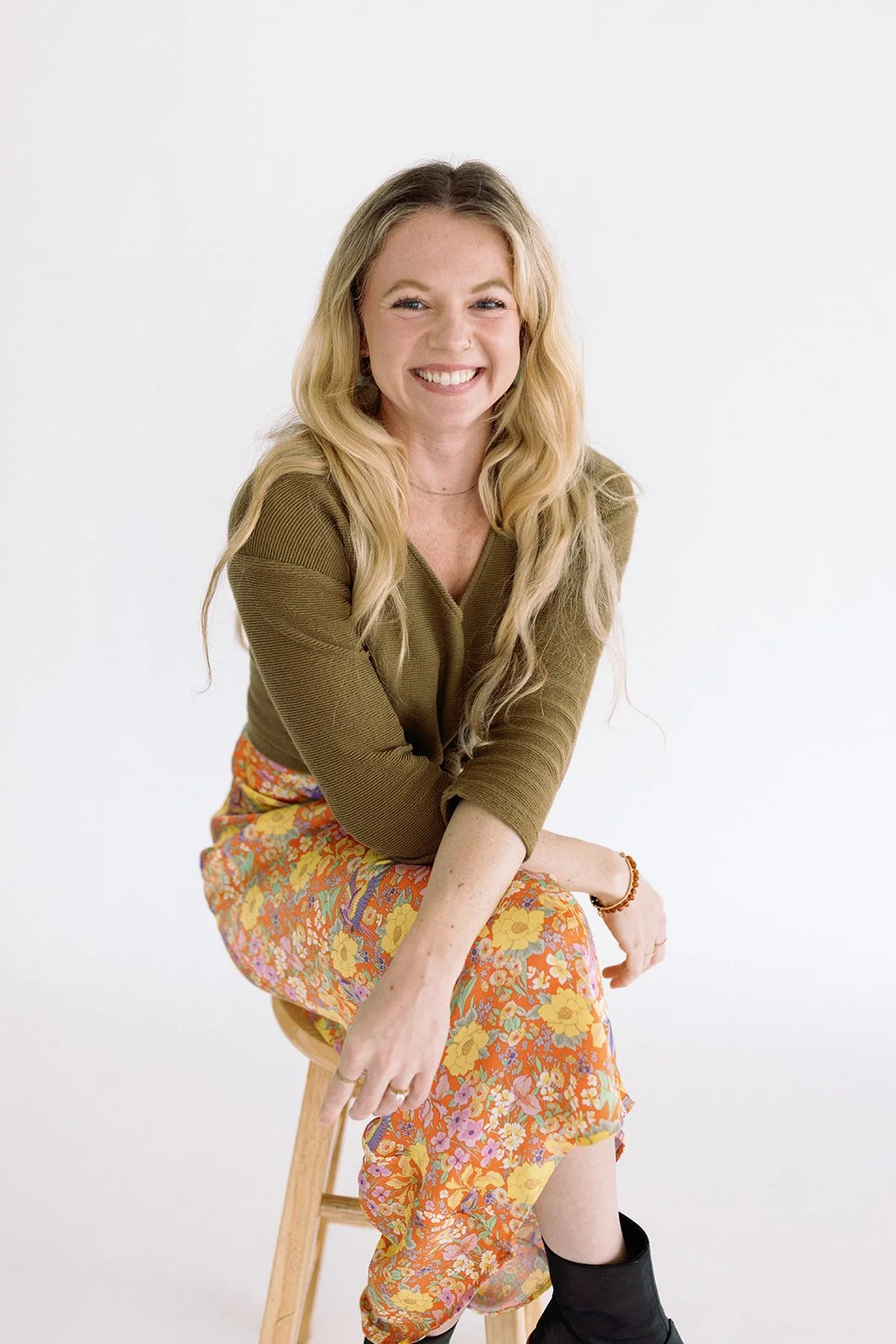 Smiling woman with long blonde hair sitting on a wooden stool in a white studio, wearing an olive top and floral skirt