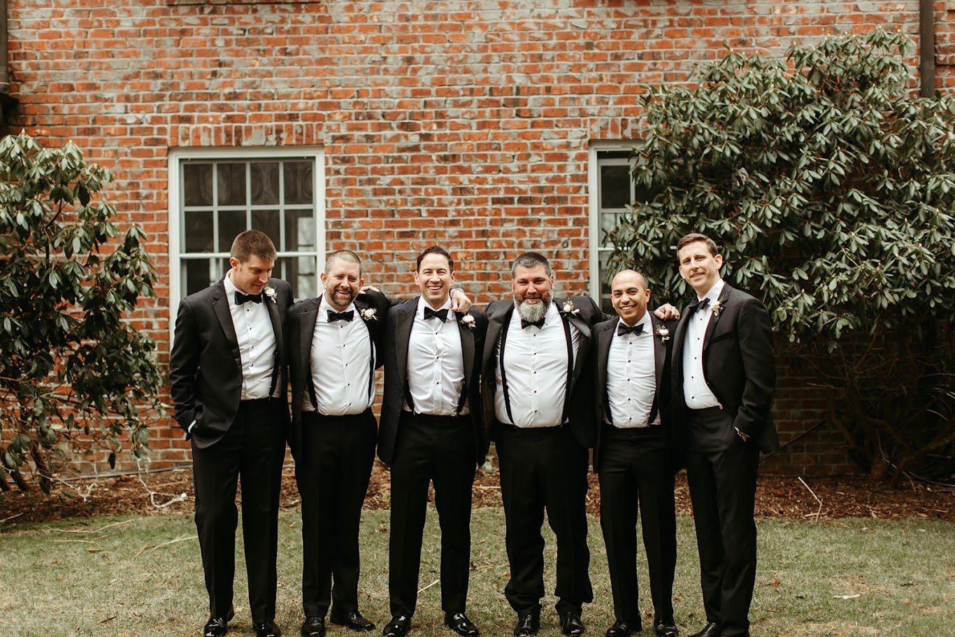 Groomsmen standing together outdoors in black tuxedos in front of a brick wall.