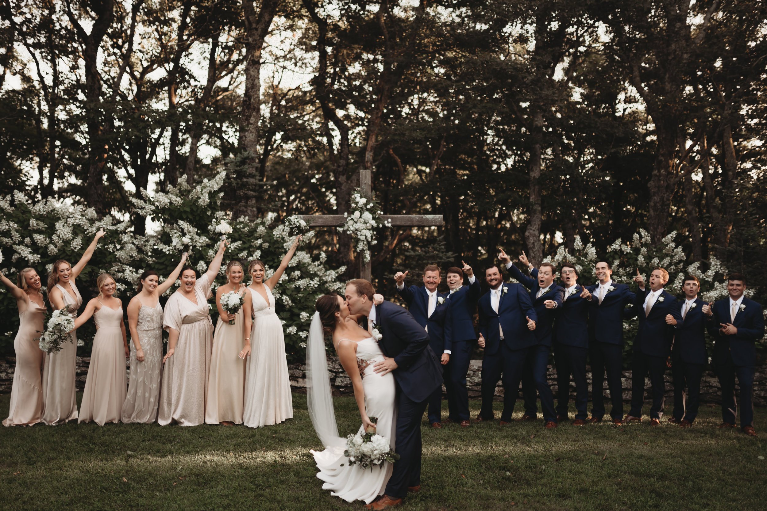 Bride and groom sharing a dip kiss with the full wedding party cheering behind them outdoors