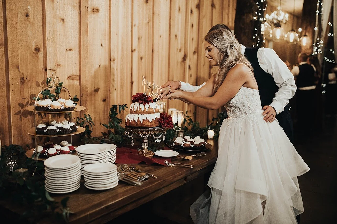 Bride cuts the wedding cake on a dessert table with cupcakes and plates, while the groom stands behind her in a rustic venue.