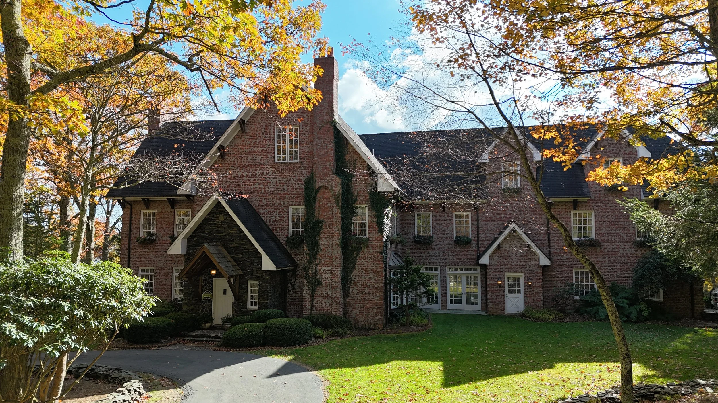 Brick manor front facade framed by golden fall branches, showing the entryway, windows, and lawn in sunlight.