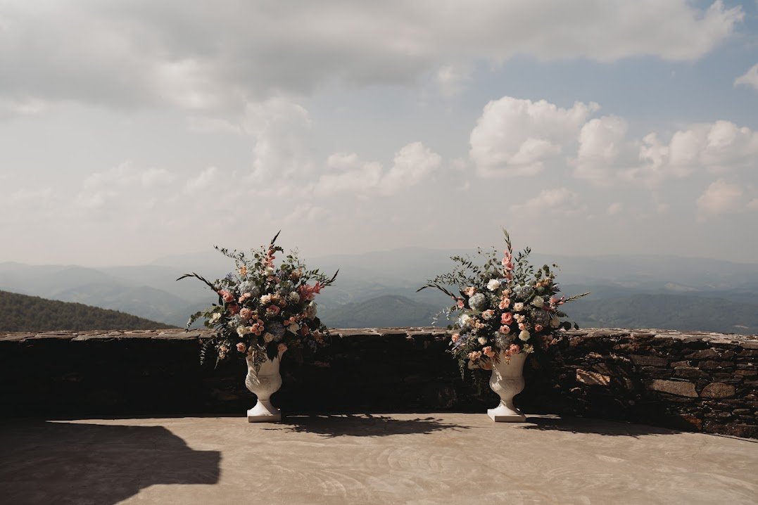 Two large floral urn arrangements on a stone terrace wall with layered mountain views in the background