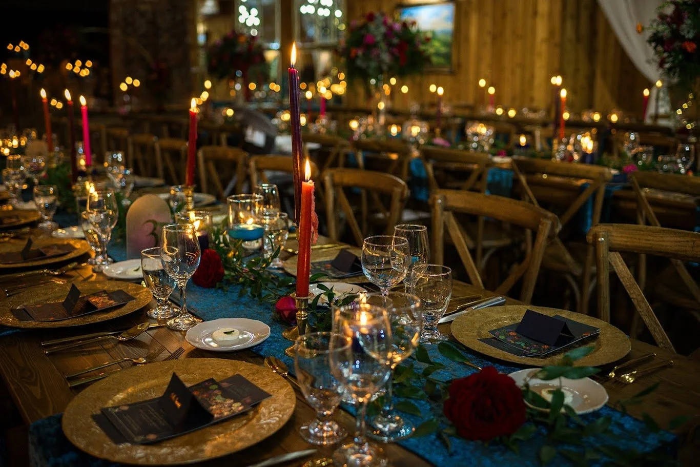 Reception table details with candles, glassware, gold chargers, and floral centerpiece in a dimly lit barn venue
