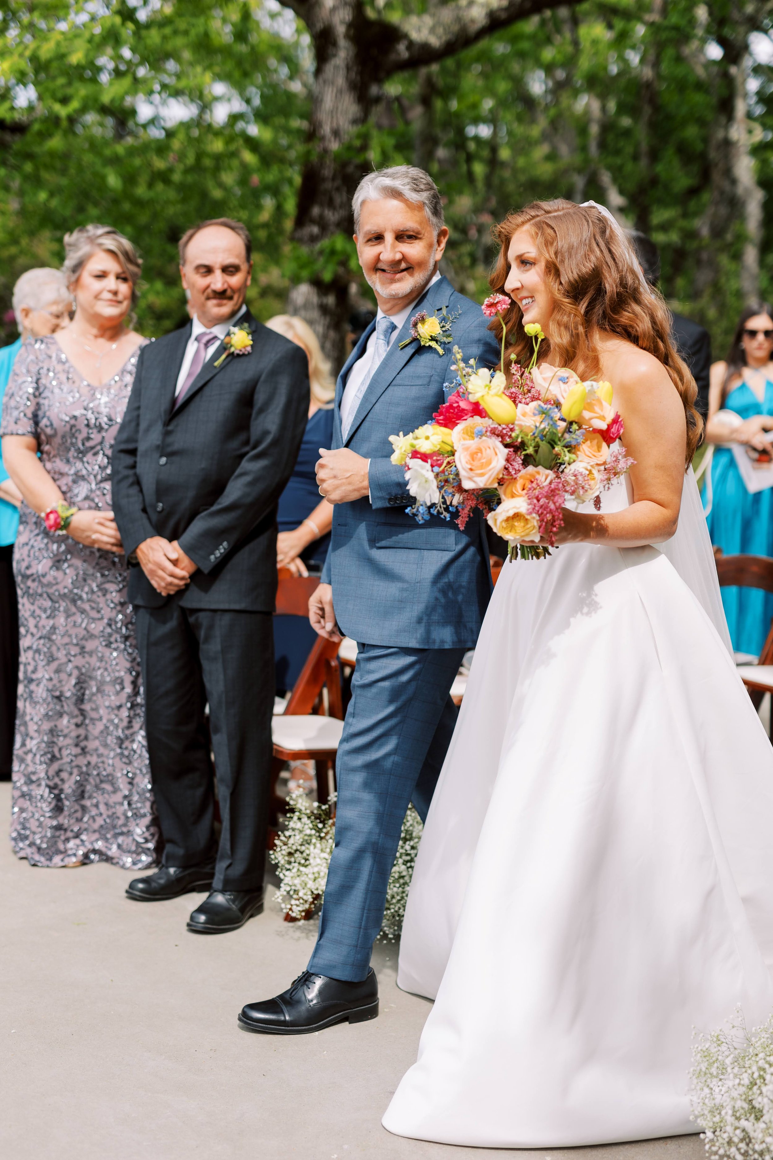 Bride walking down the aisle with her father at an outdoor ceremony, holding a colorful bouquet as guests look on.