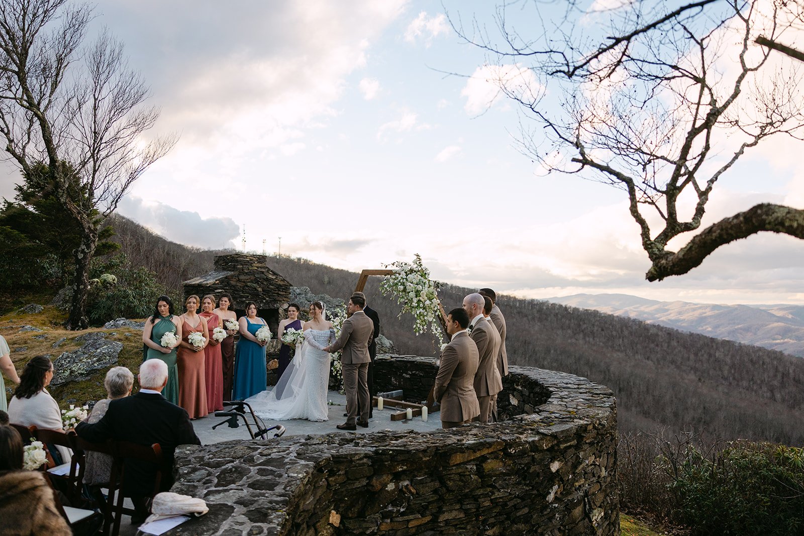 Outdoor wedding ceremony on a stone terrace overlooking mountains with the couple at the altar and guests seated nearby.