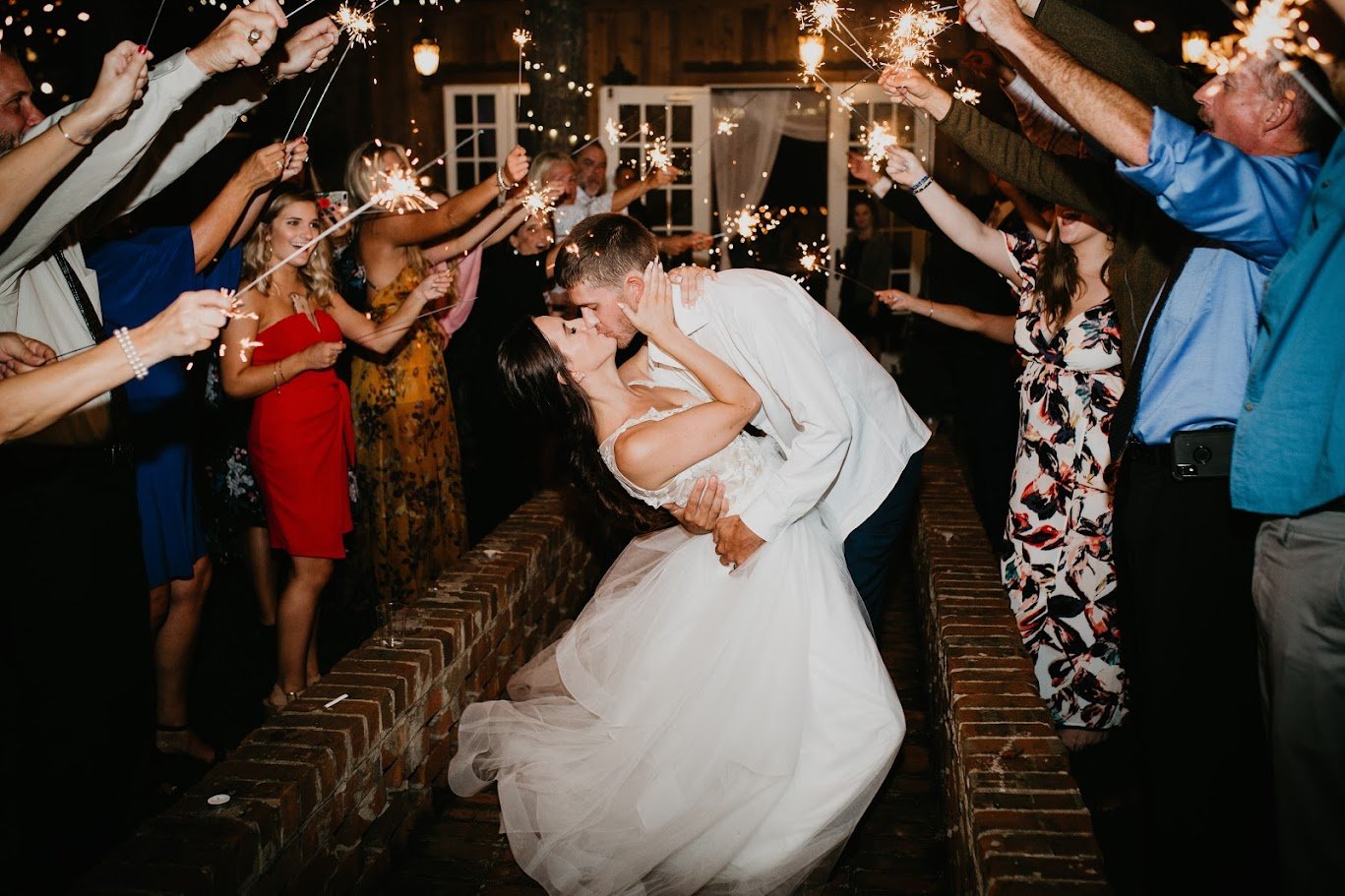 Newlyweds kissing during a sparkler exit surrounded by cheering wedding guests at night.