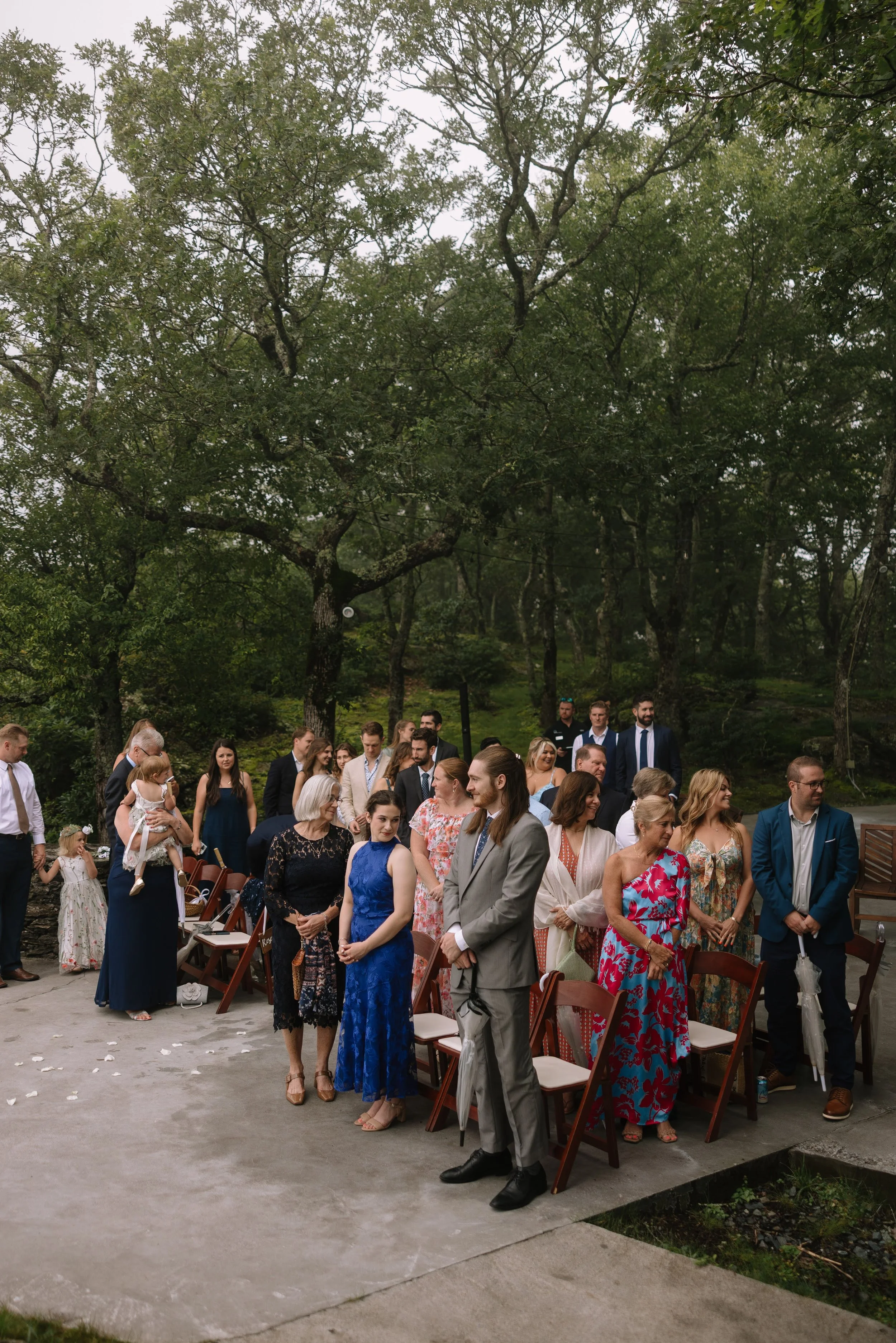 Wedding guests standing at an outdoor ceremony surrounded by trees at a wooded venue.