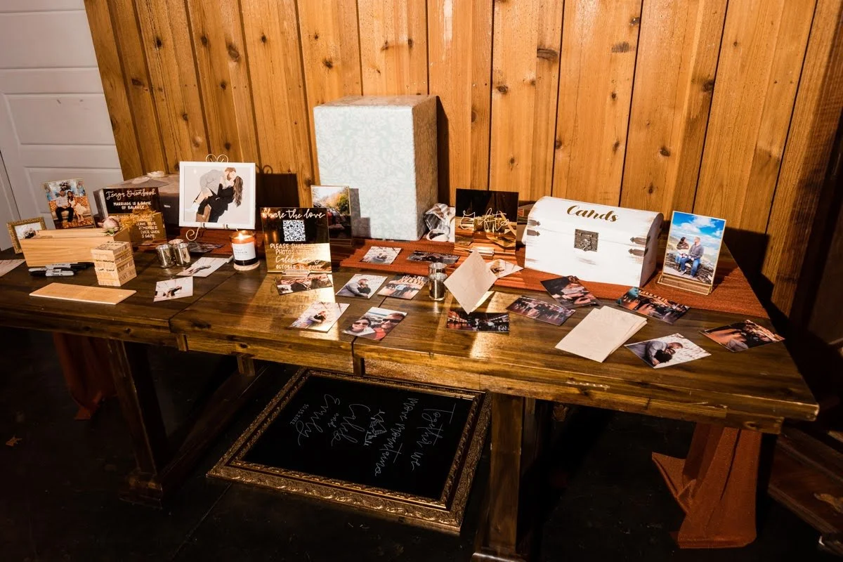 Rustic wedding guestbook table with photos, cards, and small decor items arranged across a wooden table against a wood-paneled wall.