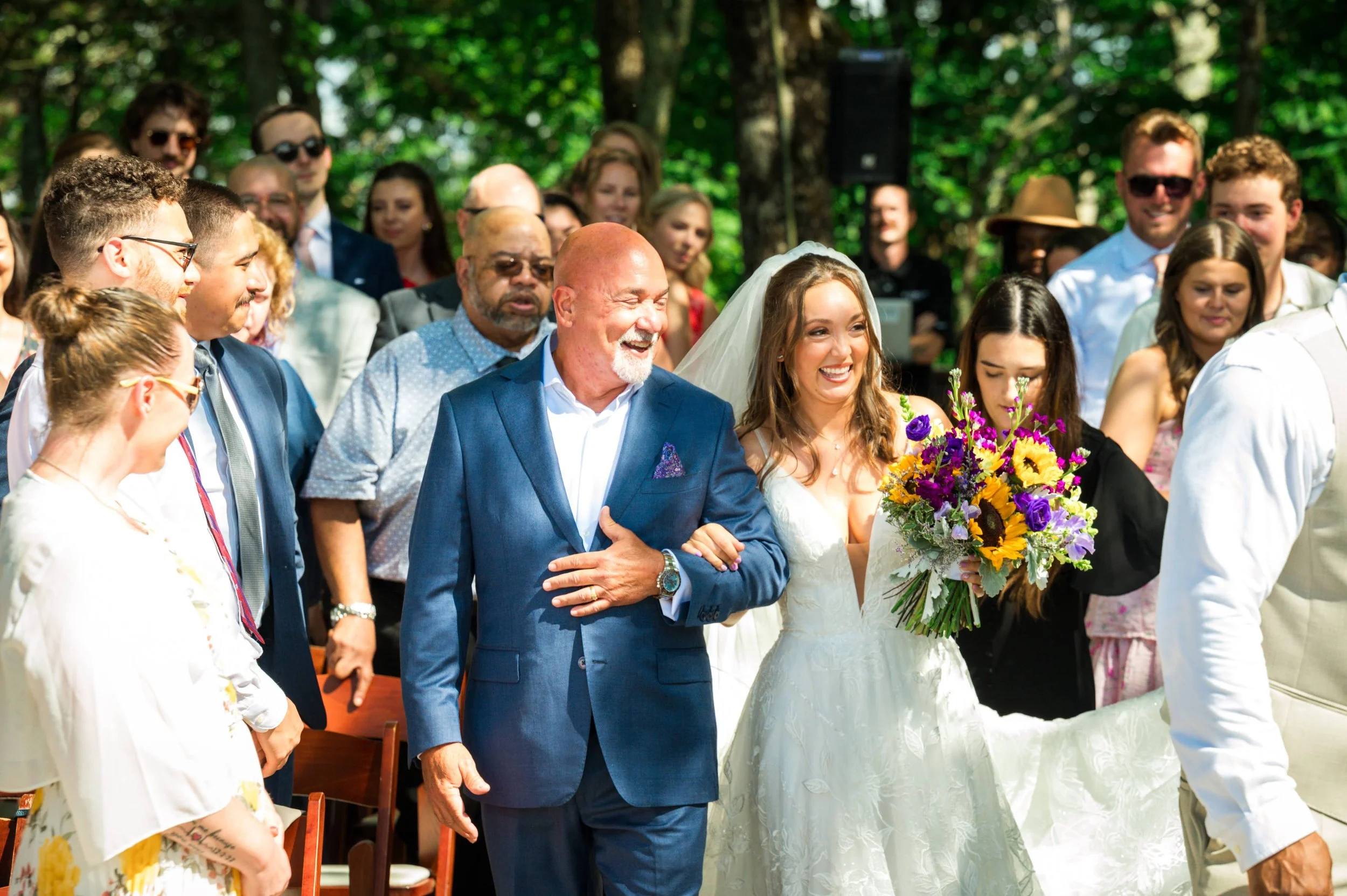Smiling bride walking down the aisle with a family member at an outdoor wedding surrounded by guests and trees