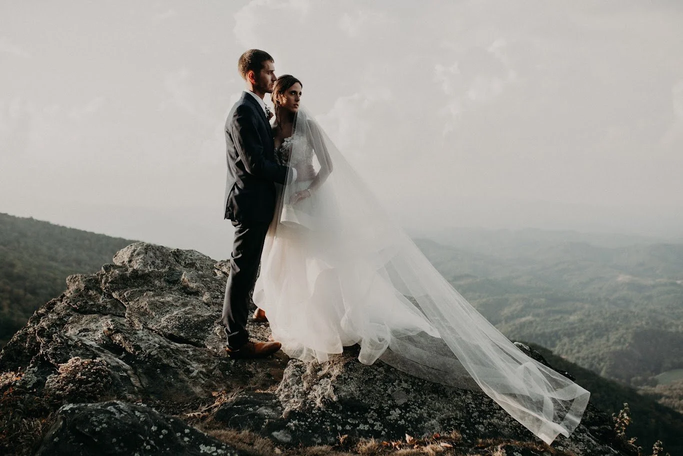 Bride and groom standing on rocky mountain terrain with a long veil and scenic valley views.