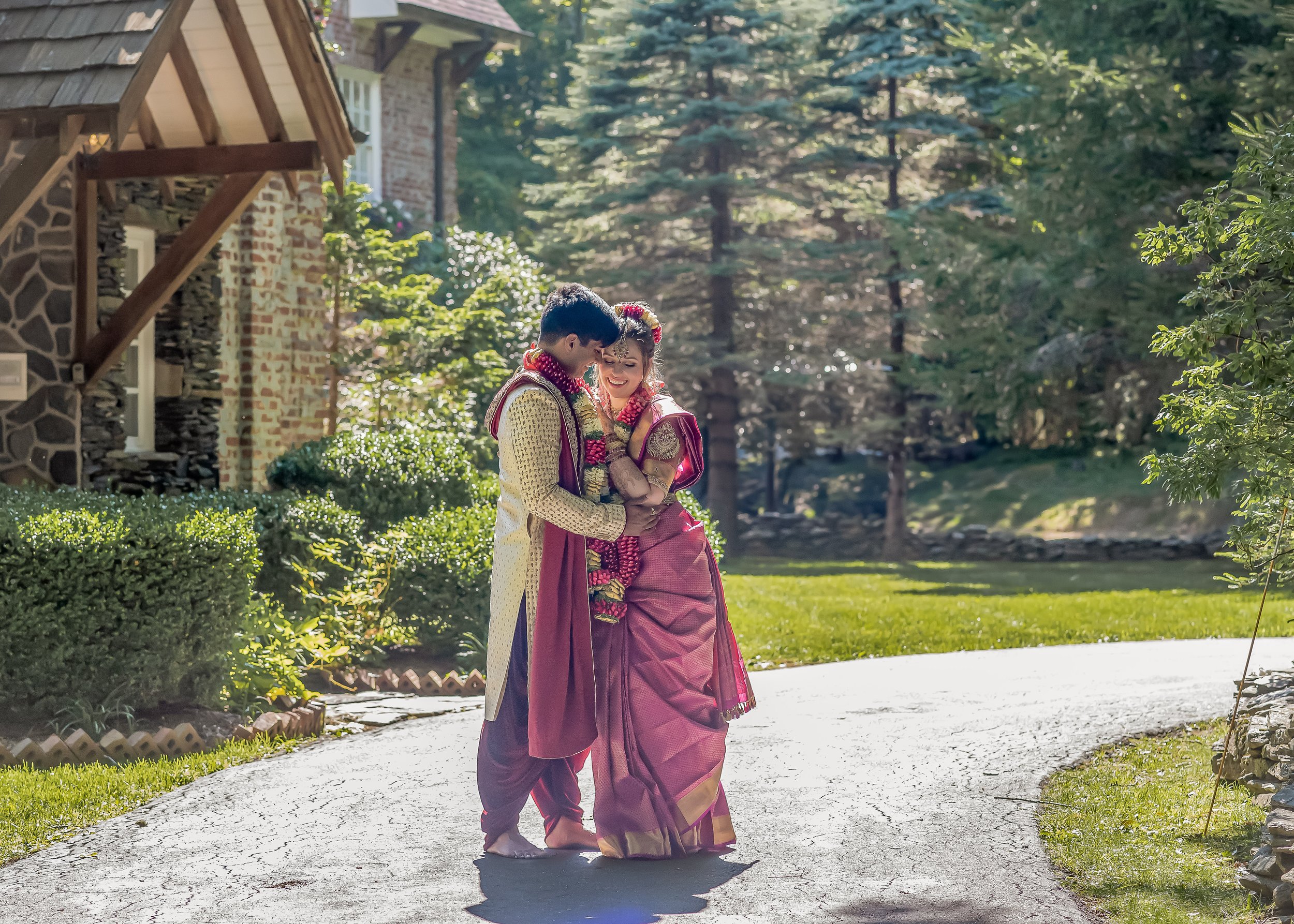 South Asian wedding couple walking together in traditional attire with flower garlands in a sunny garden.