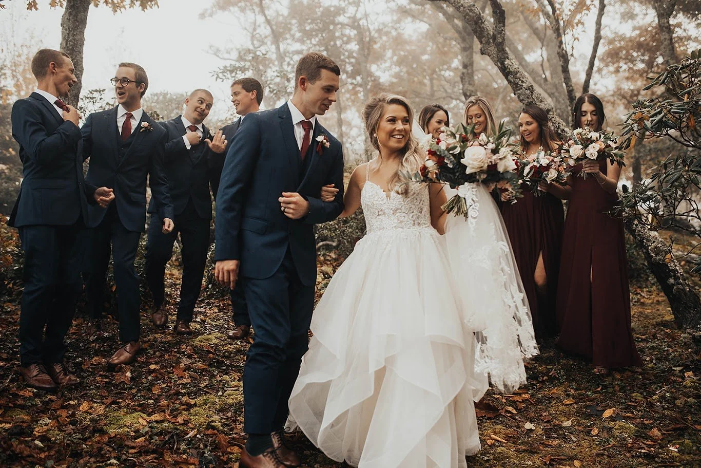 Bride and groom walking with the wedding party in the woods, bridesmaids in burgundy and groomsmen in navy suits.