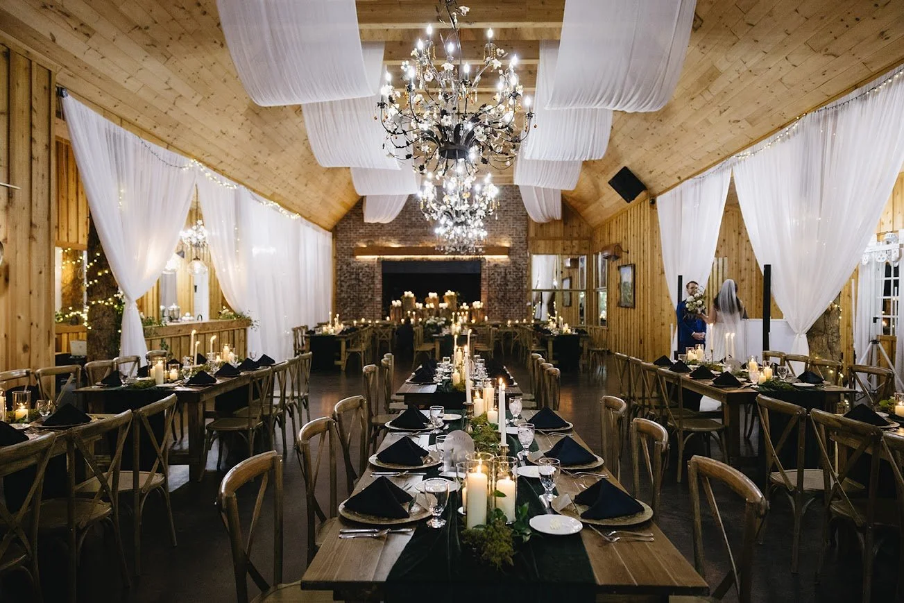 Long wooden reception tables set with black napkins and white candles under elegant chandeliers and fabric draping.