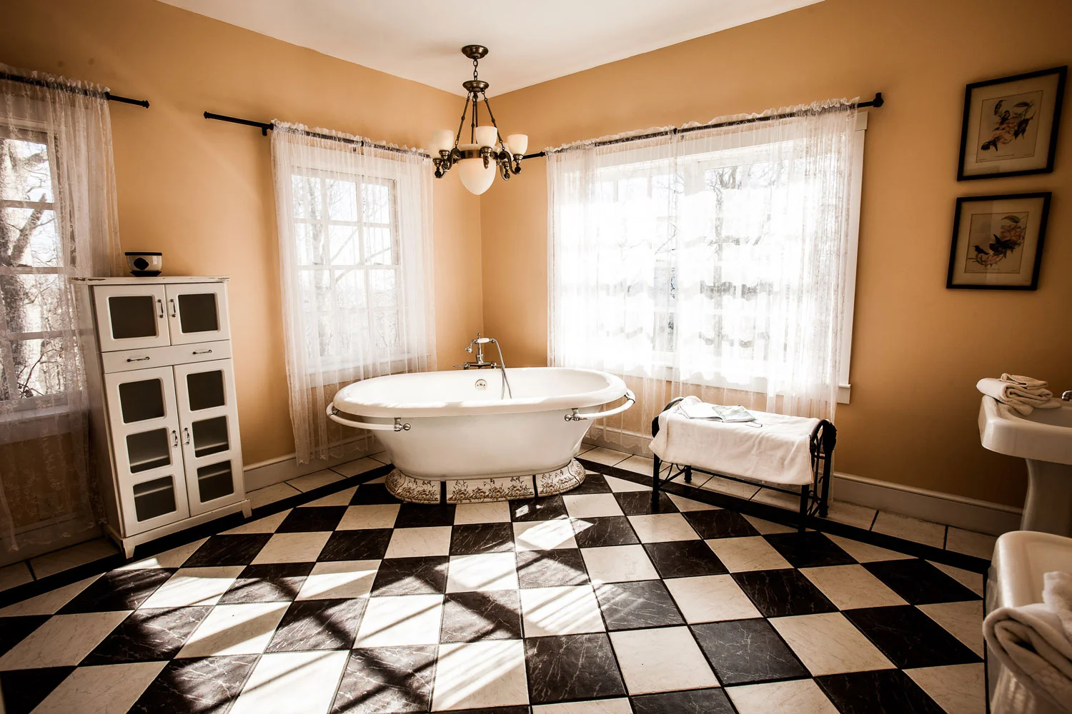 A soaking bathtub surrounded by natural light, large windows, and a hanging chandelier. His and hers sinks are against the opposite wall of the bathtub. This bathroom is part of the Versace suite.