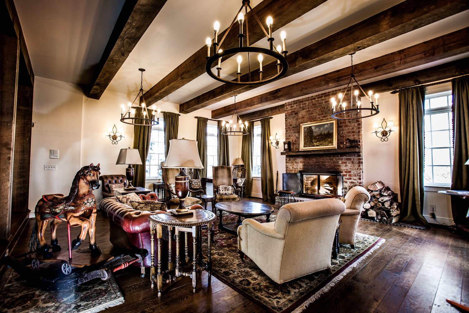  A living room with a large fireplace, hanging chandeliers, numerous cushion chairs and a red, leather couch accompanied by an antique rocking horse. 