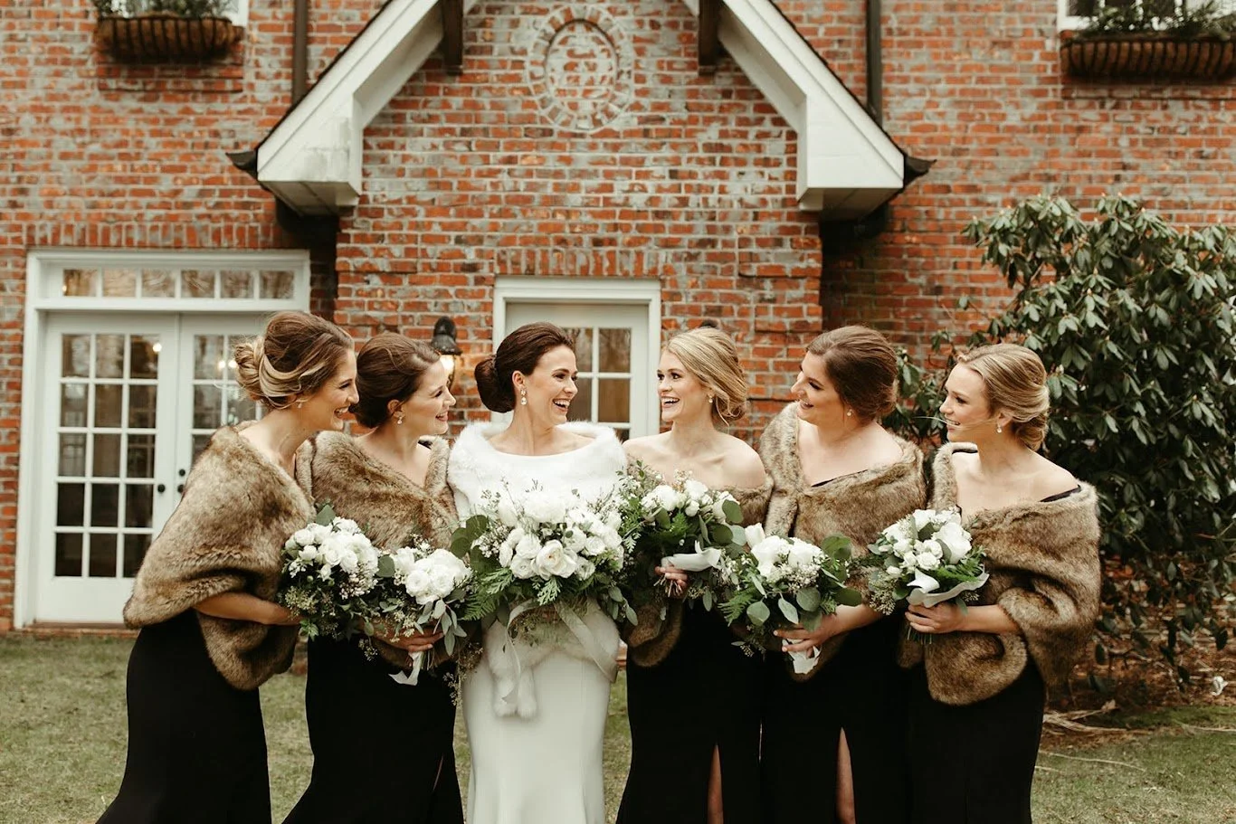 Bride with bridesmaids wearing fur wraps holding white bouquets outside a brick venue.