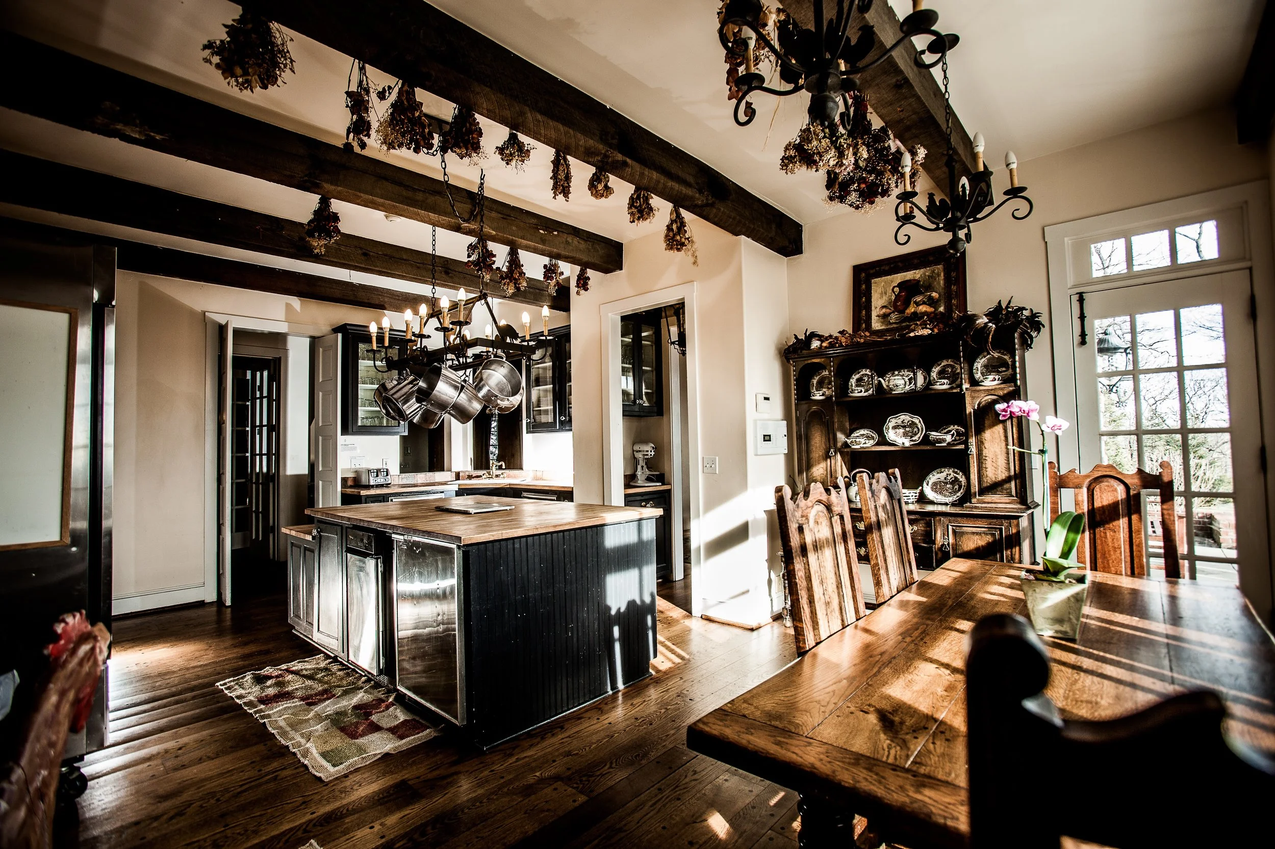 A large, sun soaked kitchen with an island and wooden breakfast table. Dried bouquets from brides hangs from the ceiling. The kitchen has an industrial fridge, stove, and other kitchen appliances.