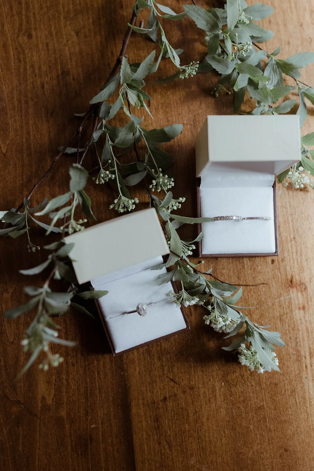 Wedding rings displayed in white ring boxes with eucalyptus greenery on a wooden surface.