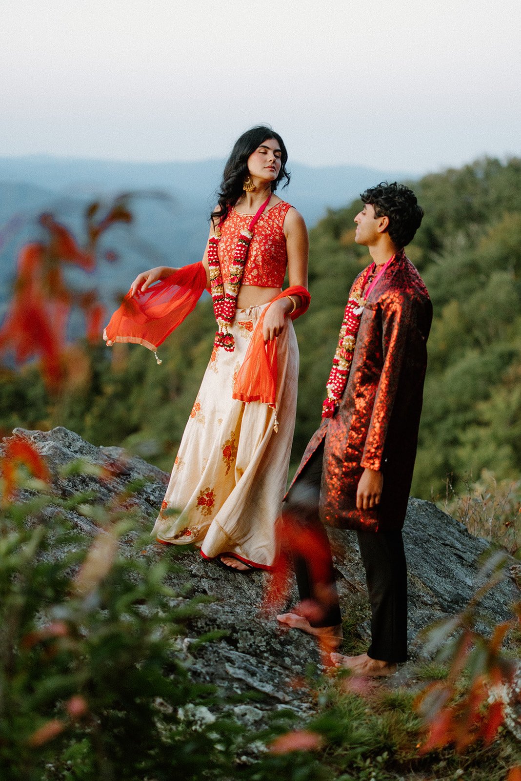 Indian couple posing on a rocky cliff, bride wearing an orange dupatta and floral garland