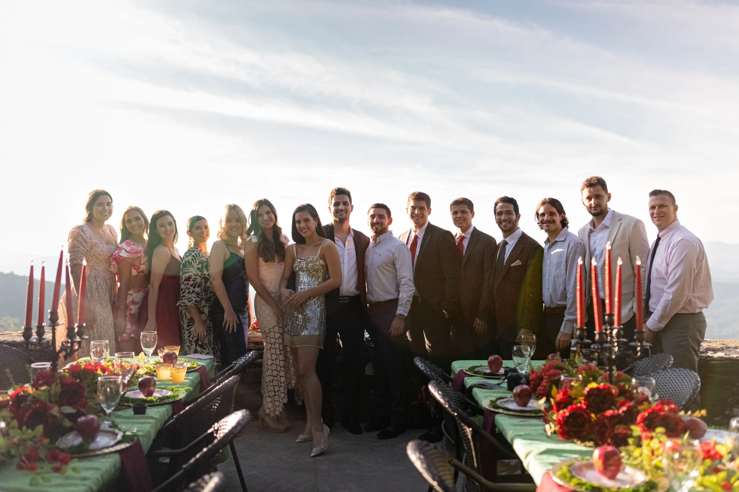 Welcome party guests posing between colorful outdoor dinner tables with red candles, florals, and mountain views.