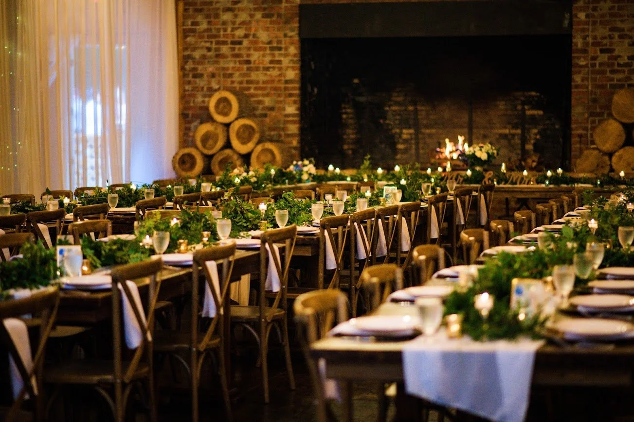 Long wooden tables decorated with greenery in a barn reception hall featuring a large brick fireplace.