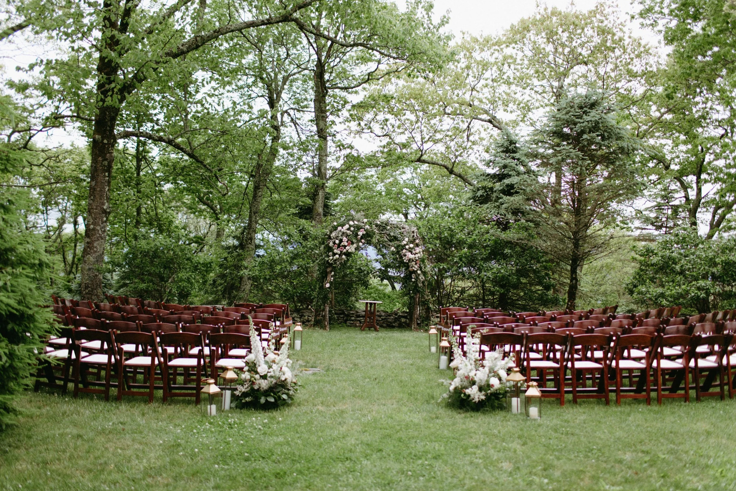 Outdoor ceremony setup on grass with rows of wooden chairs facing a floral arch in a wooded garden.