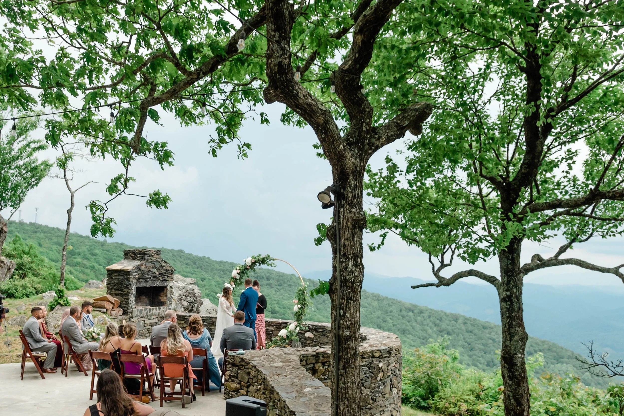 Outdoor mountain wedding ceremony beneath a large tree with guests seated near a scenic overlook.