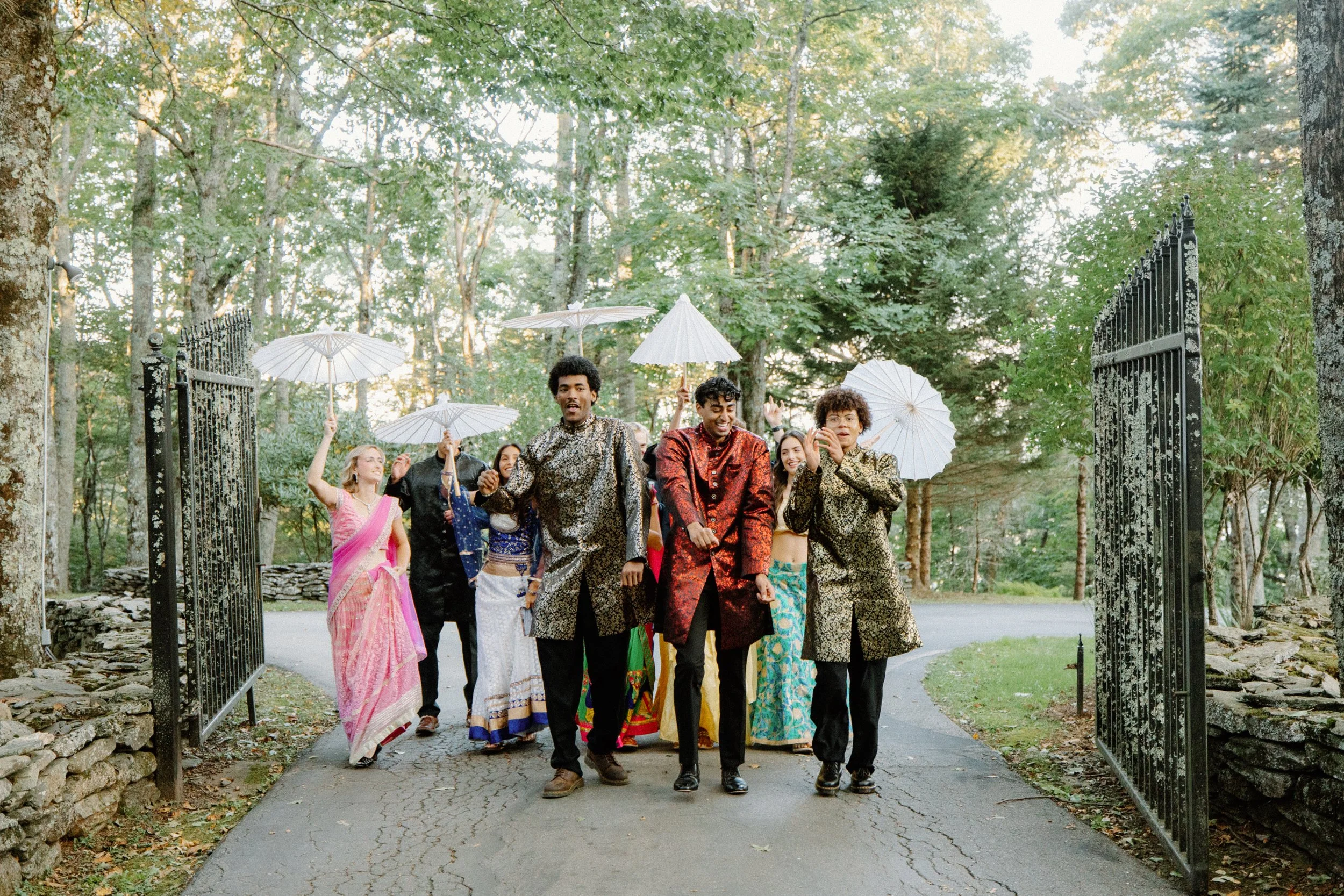 Wedding party in colorful South Asian outfits walks through open iron gates, holding white parasols on a tree-lined drive.
