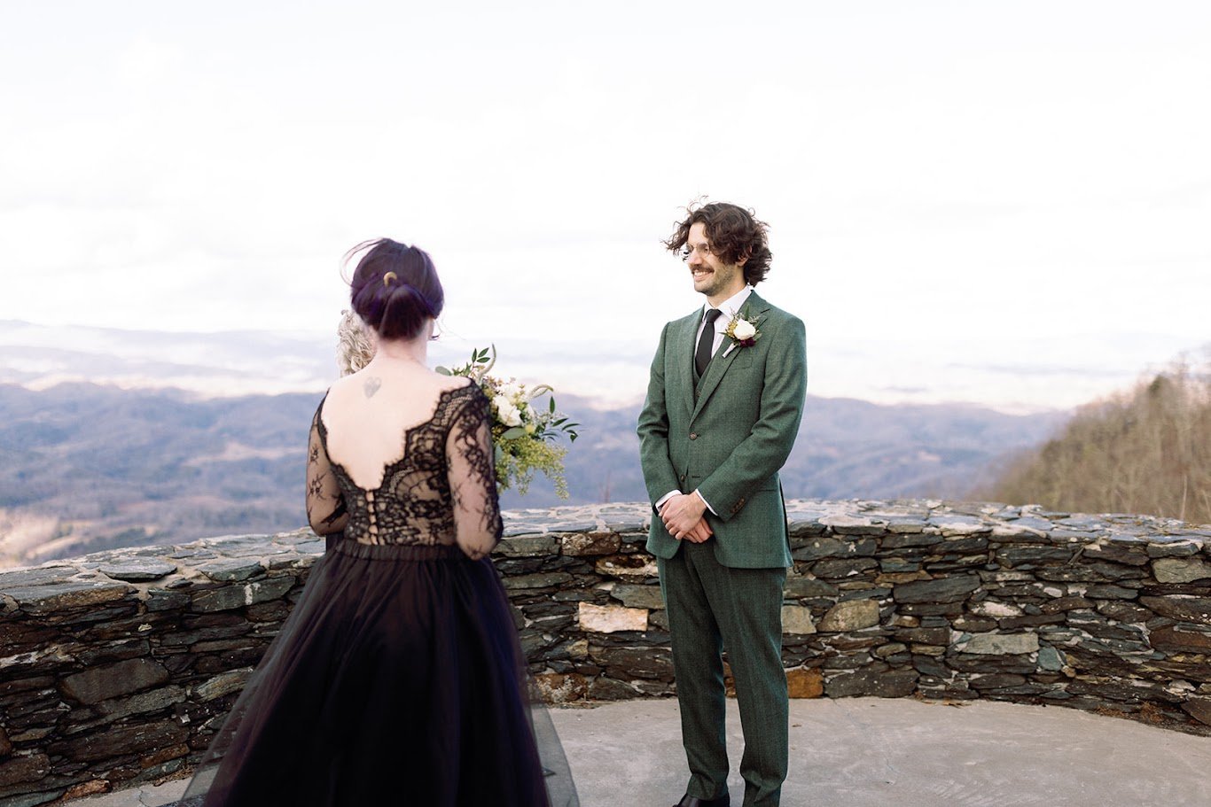A bride in a black lace dress and a groom in a green suit share a first look on a mountain overlook in North Carolina.