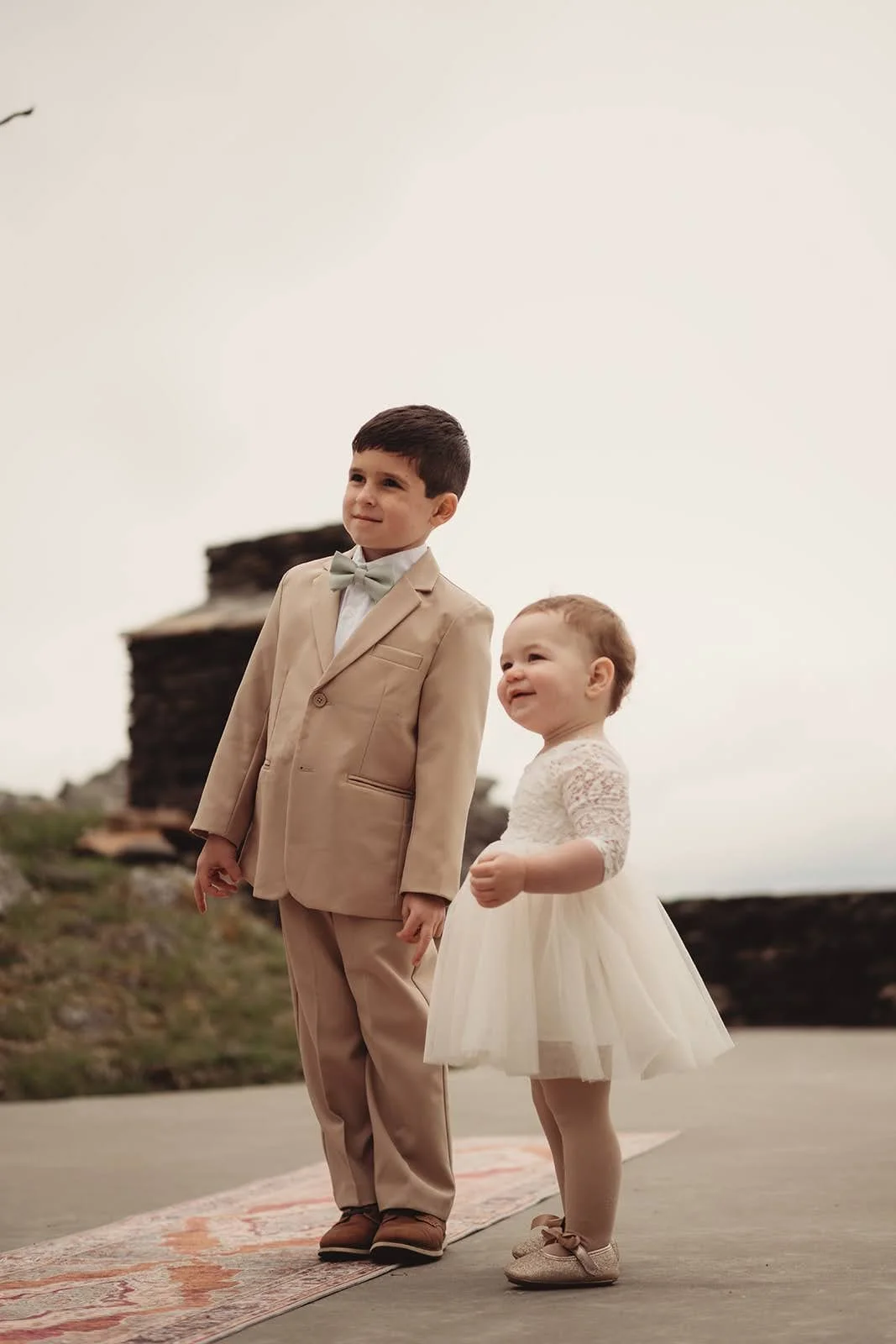 Young ring bearer in a tan suit and flower girl in a white dress stand together on a stone terrace.