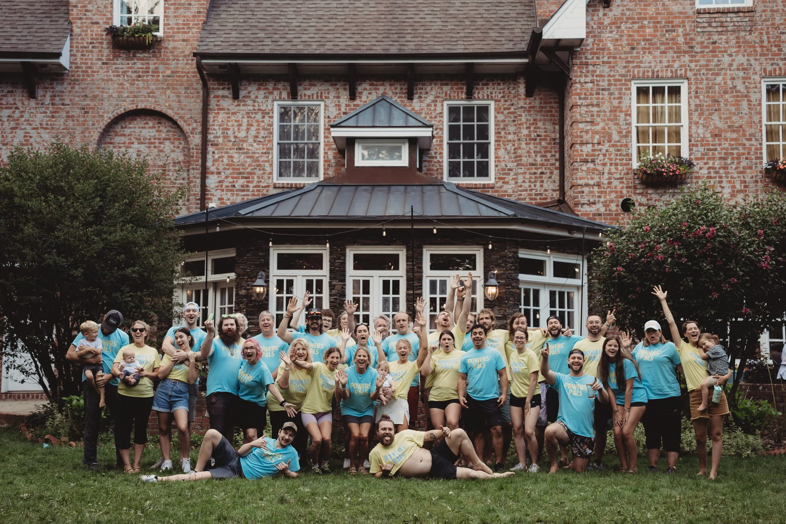 Large group photo on the lawn in front of a brick estate building during a wedding weekend