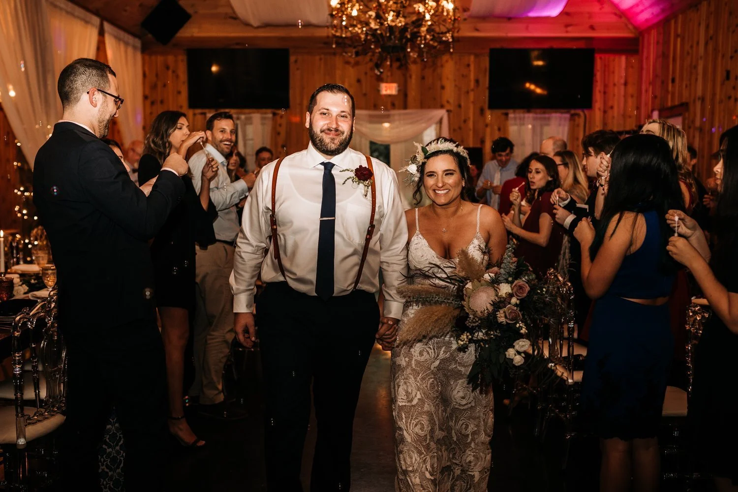 A smiling bride and groom walk down an aisle indoors, holding hands, as wedding guests blow bubbles around them.